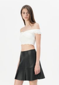 Balts off-the-shoulder top ar viļņotām malām, apvienots ar melnu ādas platmalu svārku, izceļot gludas tekstūras un pieguļošas formas.