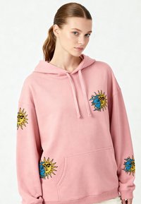 Rosa hoodie med sol- och månbroderi på ärmarna och frontfickan. Tillverkad av mjukt tyg med en avslappnad passform och dragsnodd i huvan.