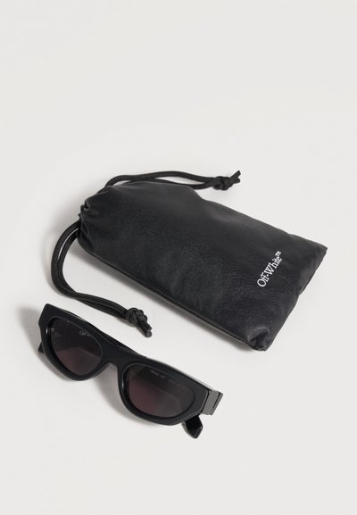 Lunettes de soleil noires style œil de chat avec verres foncés à côté d'une pochette noire à cordon de marque « Off-White » sur un fond clair.