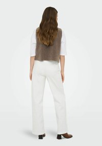 Femme aux longs cheveux ondulés portant un gilet sans manches marron texturé par-dessus une chemise blanche, un pantalon blanc et des bottes à talons marron, debout de dos.