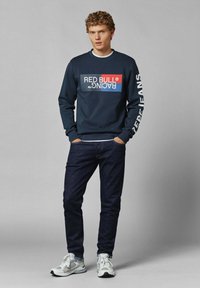 Sudadera azul marino con un gran logo en rojo y azul de "RED BULL RACING", combinada con unos vaqueros oscuros y zapatillas gris con detalles en blanco.