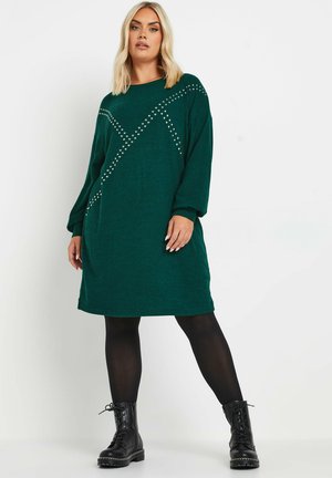 Vestido de punto verde de gran tamaño con mangas largas, que presenta acentos de tachuelas plateadas en un patrón geométrico, combinado con leggings negros y botas con cordones.