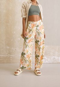 Personne portant un pantalon ample à fleurs, un crop top vert, une chemise beige ouverte, des sandales et un collier de perles, debout contre un mur texturé.