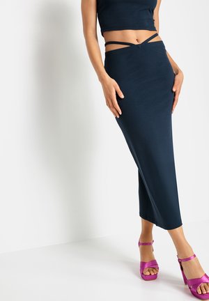 Marineblauwe geribbelde gebreide rok met een hoge taille en uitsnijdingen bij de bandjes, gecombineerd met fuchsia sandaaltjes op een witte achtergrond.