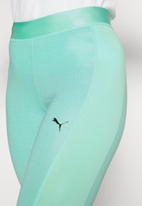 Zbliżenie na jasnoniebieskie legginsy sportowe z teksturowanym wzorem i czarnym logo Pumy na udzie, noszone z białą bluzką.