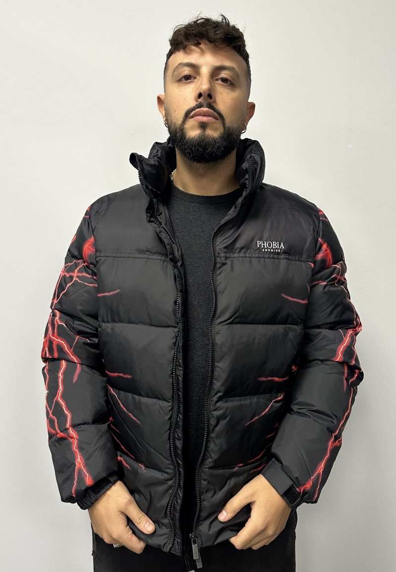 Giacca a vento nera con grafiche di fulmini rossi, chiusura con zip, collo alto e polsini elasticizzati. Il materiale appare lucido e texture.