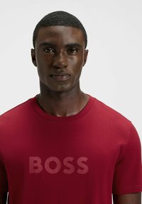 Rotes Baumwoll-T-Shirt mit Rundhalsausschnitt und einem großen, halb-transparenten "BOSS"-Logo auf der Brust, glatter Textur und kurzen Ärmeln.
