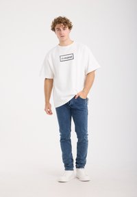 Witte oversized T-shirt met een zwart rechthoekig logo op de borst, gecombineerd met blauwe jeans en witte sneakers, staand tegen een effen achtergrond.