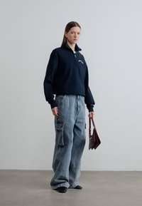 Σκούρα μπλε φούτερ με φερμουάρ, συνδυασμένο με άνετο γκρίζο τζιν παντελόνι cargo με τσέπες, κρατώντας μια τσαντάκι σε καφέ-κόκκινη απόχρωση. Απαλές υφές, χαλαρή εφαρμογή.