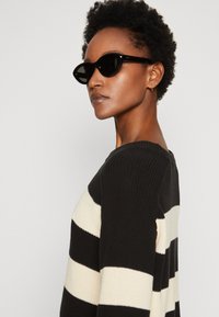 Maglione a righe bianche e nere con una texture a coste, dotato di scollo rotondo e maniche lunghe. Modella che indossa occhiali da sole a forma di occhio di gatto neri.