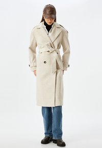 Abrigo beige de estilo envolvente con cinturón de lazada, grandes botones y hombreras; se lleva sobre un suéter de cuello alto negro y jeans azules.