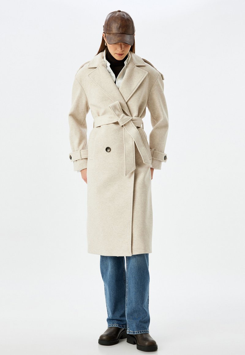 Beige Wickelmantel mit Bindegürtel, großen Knöpfen und Schulterklappen; getragen über einem schwarzen Rollkragenpullover und blauen Jeans.