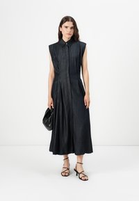 Mørk denim ermeløs kjole med knappestolpe, strukturert krage, folder og en flytende silhuett. Modellen holder en liten svart veske.