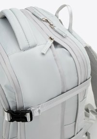 Mochila de color gris claro con un diseño acolchado, que cuenta con cremalleras dobles, correas ajustables y un cierre de hebilla para mayor seguridad.