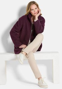 Fleece-Überziehjacke in Dunkelpurpur mit strukturiertem Finish, kombiniert mit cremefarbenen Hosen und weißen Sneakern. Einfaches Design mit Stehkragen.