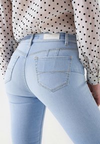 Los jeans skinny azul claro tienen una cintura alta, cinco bolsillos y detalles de costura sutiles. La tela tiene una textura de denim suave.