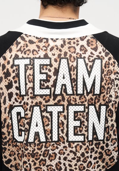 Άτομο που φοράει μπουφάν με σχέδιο λεοπάρδαλης με μαύρα μανίκια και λευκό κείμενο "TEAM CATEN" στην πλάτη.
