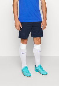 Pantalones cortos deportivos de color navy con detalle del logo, acompañados de calcetas blancas hasta la rodilla con acentos morados y botas de fútbol turquesa con tacos amarillos.