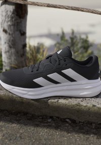 Baskets de sport noir et blanc avec dessus en mesh et trois bandes blanches, placées sur une surface en pierre à l’extérieur.