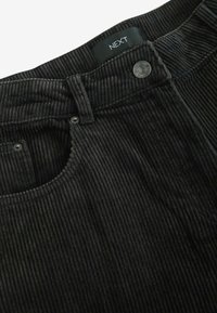 Pantalons en velours côtelé noir avec texture en côtes verticales, fermeture à bouton métallique et détails de poche. Étiquette de marque visible à l'intérieur de la taille.