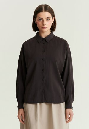 Woman with short brown hair wearing a loose black button-up shirt and a light beige skirt, standing against a plain light background.