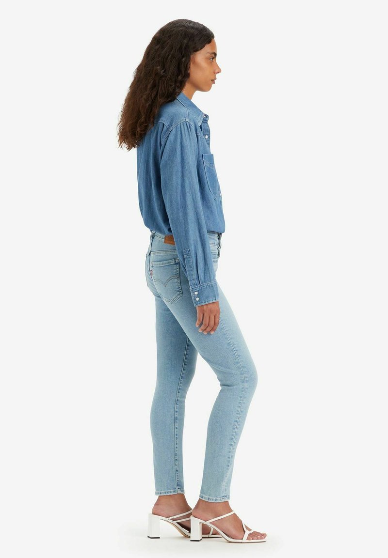 Camisa de mezclilla en azul claro con mangas largas y bolsillo en el pecho. Jeans ajustados en azul claro combinados con sandalias de tacón blanco. Look limpio y elegante.