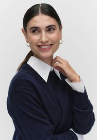 Donkere marineblauwe trui met een witte blouse. Het model heeft lang, steil donker haar en draagt kleine ringoorbellen.