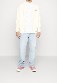 Light yellow cardigan with front buttons, worn over a white shirt. Paired with light blue jeans and red sneakers with white soles.