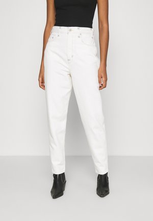 Persoon draagt een witte, hooggetailleerde jeans gecombineerd met zwarte enkellaarsjes en een mouwloze zwarte top tegen een witte achtergrond.