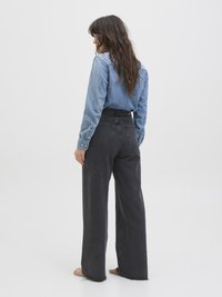 Kvinne med langt brunt hår står barføtt med siden til, iført en lyseblå denimskjorte og brede, svarte jeans mot en hvit bakgrunn.