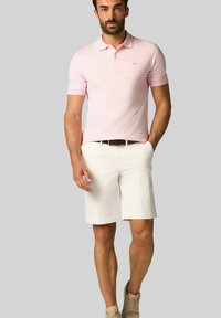 Světle růžové polo tričko s krátkým rukávem a malým modrým logem. Vytvořeno s bílými kraťasy a béžovými teniskami, stojící na neutrálním pozadí.