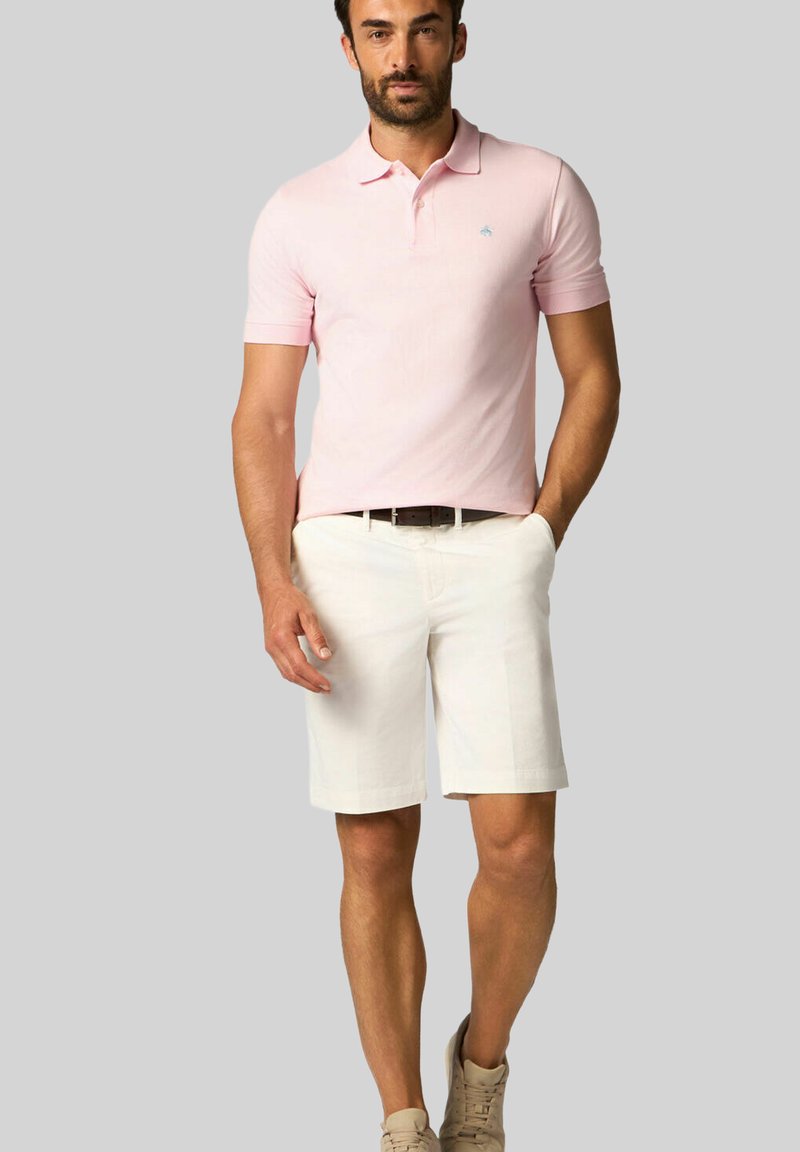 Světle růžové polo tričko s krátkým rukávem a malým modrým logem. Vytvořeno s bílými kraťasy a béžovými teniskami, stojící na neutrálním pozadí.