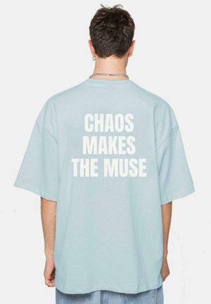Camiseta azul claro de gran tamaño hecha de algodón con el texto "EL CAOS INSPIRA" en letras blancas en negrita en la parte posterior. Mangas cortas.
