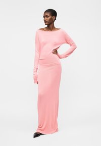 Γυναίκα που φοράει ένα μακρύ ροζ εφαρμοστό φόρεμα με μακριά μανίκια και μαύρα μυτερά παπούτσια, στέκεται με το ένα χέρι στη μέση.