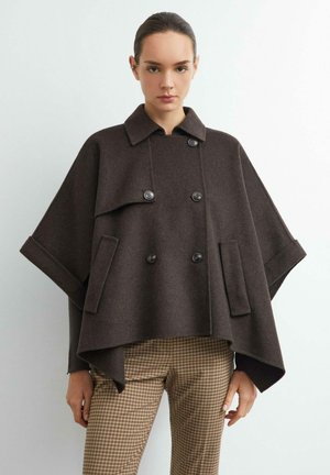 Femme aux cheveux foncés portant un manteau cape croisé marron foncé et un pantalon à carreaux beige, debout sur un fond clair uni.