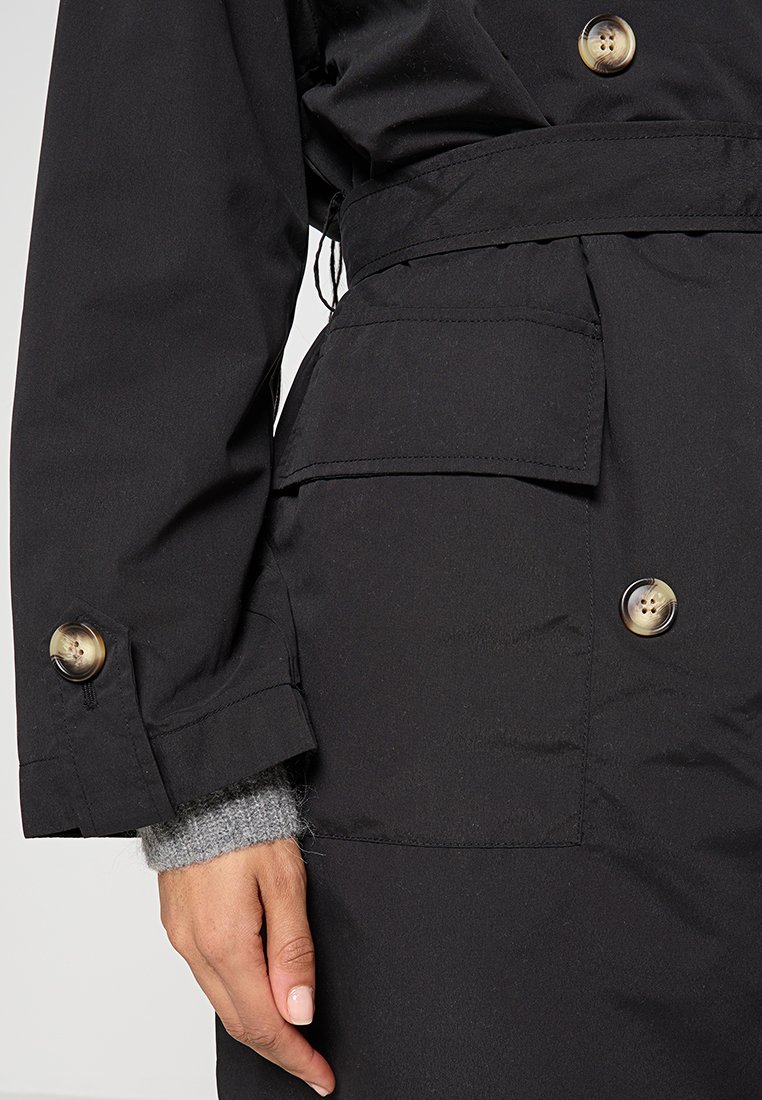 Schwarze Oberbekleidung mit Knopfverschluss, vorderen Taschen und einem taillierten Gürtel. Das Material wirkt glatt mit einem matten Finish.