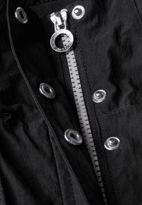Czarna tkanina kurtki z metalowym suwakiem z okrągłym uchwytem, srebrnymi zatrzaskami i teksturowanymi akcentami wzdłuż suwaka.