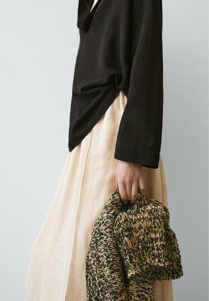 Person wearing a loose black long-sleeve top and beige skirt, holding a black and beige knitted sweater by their side against a plain background.