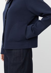 Osoba nosząca granatową zapinaną kurtkę z kieszeniami po bokach i ciemne dżinsy, z ręką włożoną w kieszeń kurtki, na jednolitym tle.