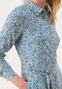 Camicia abbottonata con motivo floreale in tonalità blu e bianco. Presenta un colletto, maniche lunghe e un dettaglio legato in vita. Tessuto di cotone.