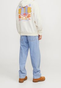 Krämfärgad huva med färggrafisk tryck på baksidan, som kombineras med ljusblå baggy jeans och bruna stövlar. Avslappnad streetwear-stil.