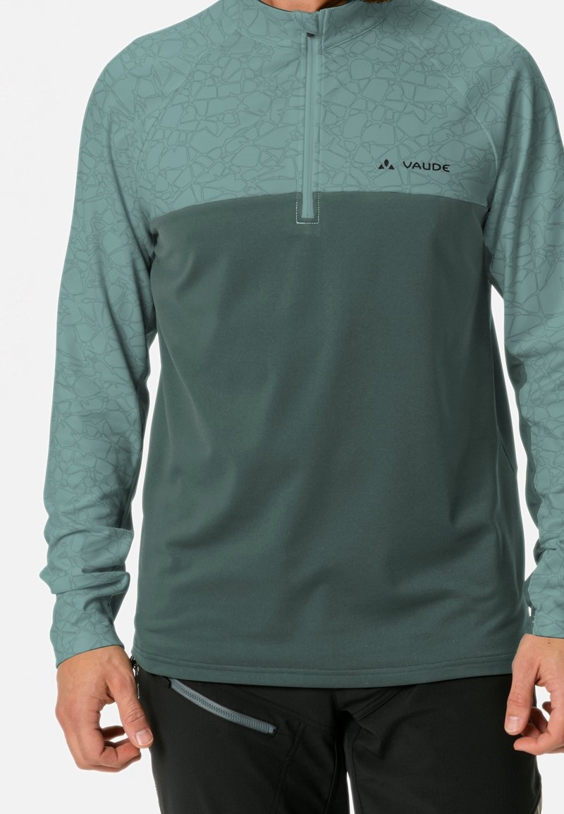 Homme portant un haut de sport à manches longues teal avec un motif géométrique sur les manches et une fermeture éclair sur la poitrine, associé à un pantalon noir avec une poche zippée.