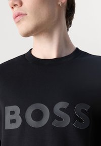 Zwarte katoenen t-shirt met ronde hals en een groot, glanzend "BOSS" logo in contrasterend materiaal, centraal op de voorkant.