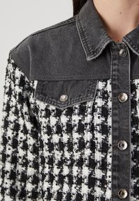 Čierno-biely kockovaný blazer so sequinmi, so tmavomodrým denimovým golierom, predným vreckom a kovovými gombíkmi na zapínanie.