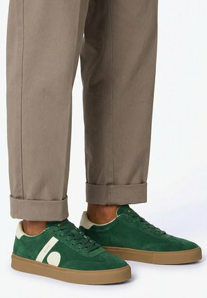 Personne portant un pantalon kaki retroussé aux chevilles et des baskets en daim vert avec des accents beiges et des semelles en gomme, se tenant sur une surface blanche.