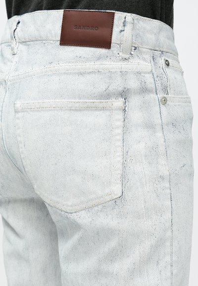 Τζιν παντελόνι από ανοιχτό μπλε τζιν ύφασμα με διακριτική φθαρμένη υφή, με καφέ δερμάτινο λογότυπο "SANDRO" στη μέση και τυπικές πίσω τσέπες.