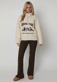 Beige fleecetröja med halvlång dragkedja, med texten "NORDIC TRAIL" och en bergsgrafik. Kombinerad med mörkbruna utsvängda byxor och beige tofflor.