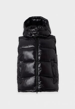 Femme portant une doudoune noire sur un pull blanc, un jean bleu à jambes larges et des chaussures noires, debout sur un fond clair uni.