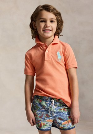 Małe dziecko o falowanych włosach, ubrane w pomarańczową koszulkę typu polo i kolorowe, wzorzyste spodenki, stojące na neutralnym tle.