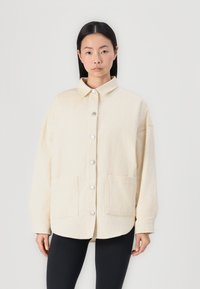 Veste en velours côtelé beige avec col, boutons-pression sur le devant et deux poches plaquées inférieures ; présente une coupe décontractée et une texture côtelée.